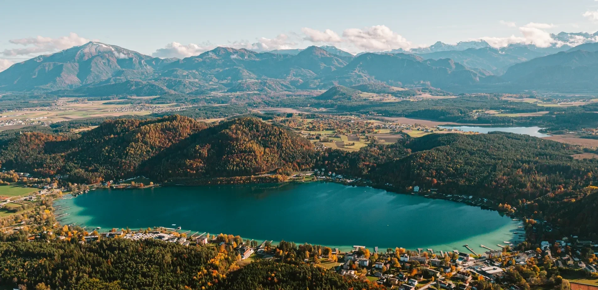 Aerial view of a lake surrounded by colorful autumn trees, rolling hills, and distant mountains under a partly cloudy sky.