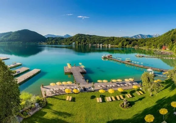 A scenic lake surrounded by lush green mountains with a clear blue sky. There is a grassy area with yellow umbrellas, lounge chairs, and a dock extending into the water.